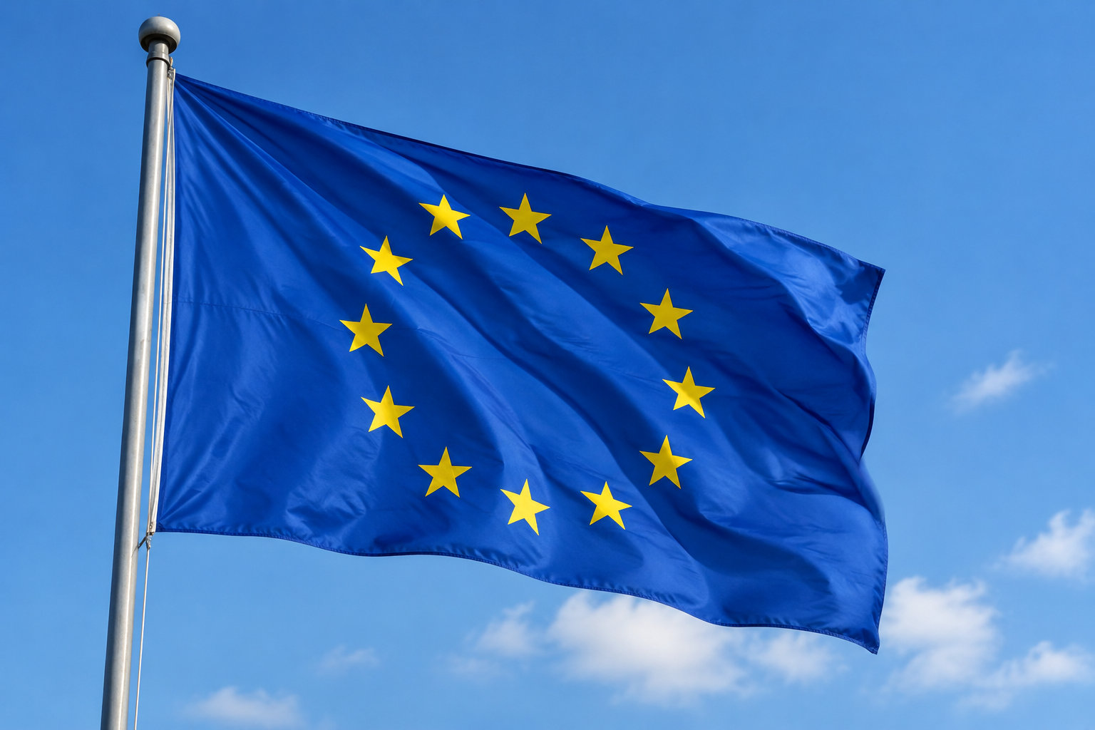 Un drapeau bleu orné de douze étoiles jaunes flotte au vent sur un mât, devant un ciel bleu nuageux.